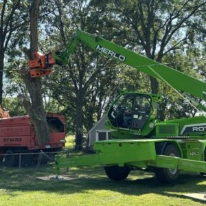 A specialized green Merlo crane and cutting attachment operates on a residential property in Olathe, KS, with the Midwest Tree crew working to remove a large tree safely.