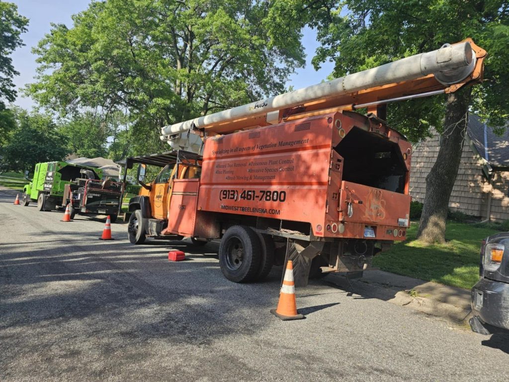 An orange Midwest Tree utility bucket truck and safety cones set up on a residential street in Lenexa, Kansas, with a green wood chipper in the background and leafy green trees overhead.